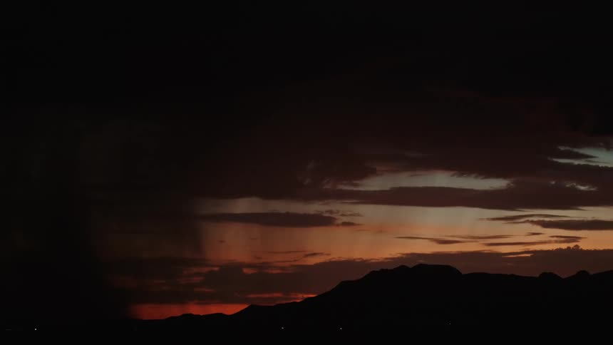 A storm with lightning bolts flashing out of the sky against an orange dusk evening with a silhouette of a mountain