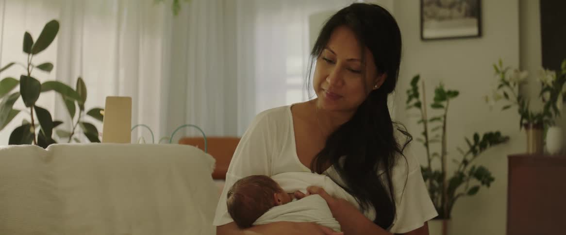 A mother nursing and rocking her newborn baby while sitting on a couch in her living room during the day
