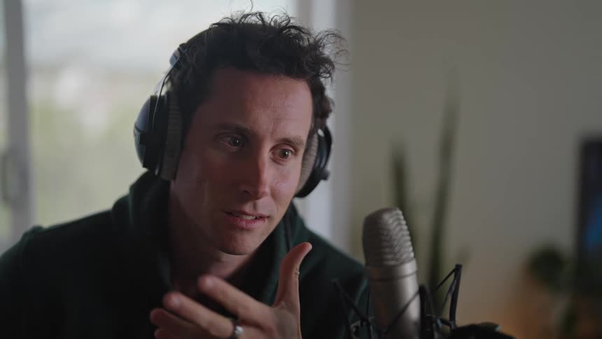Close up of a man wearing headphones speaking into a microphone in front of him while gesturing with his hand