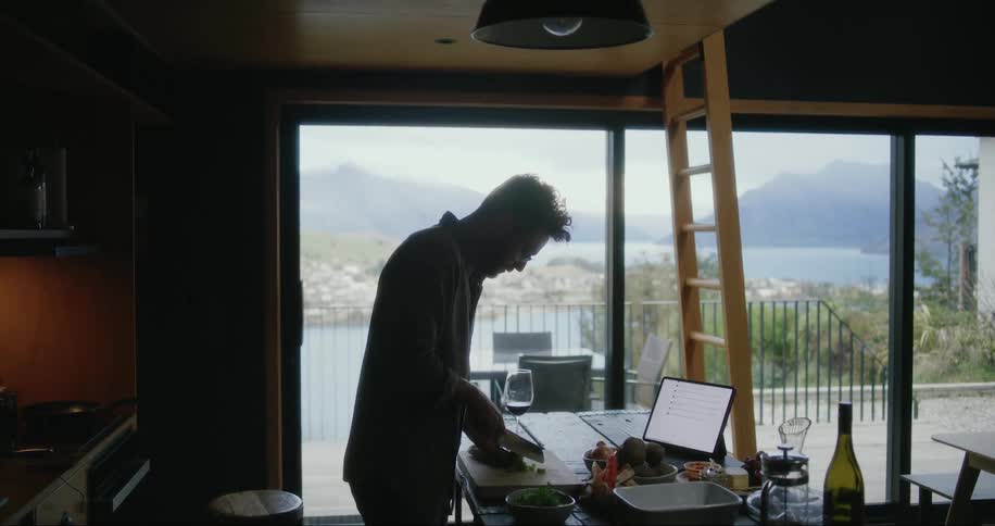 A man standing in his kitchen cutting and preparing food on the counter in front of a laptop screen in the middle of the day