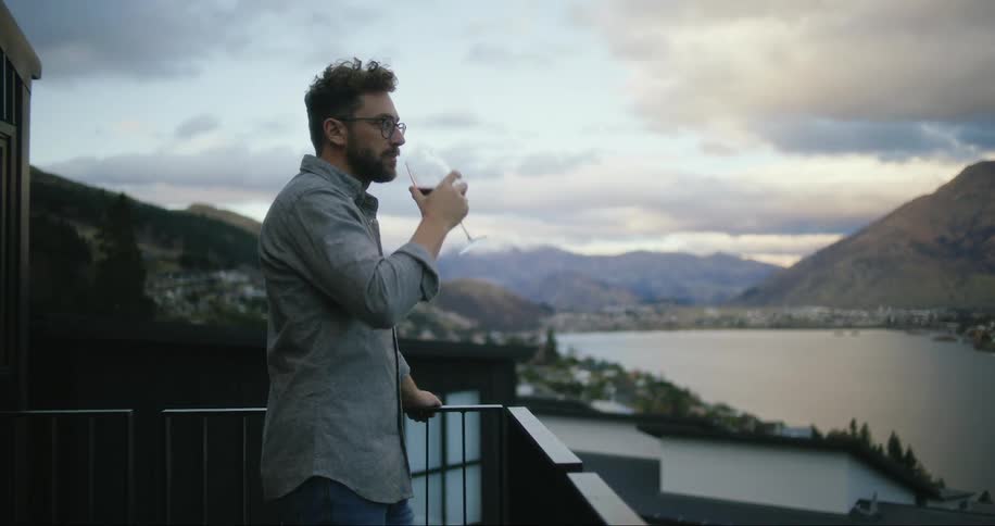 A man standing leaning against a railing while drinking a glass of wine looking out over a body of water surrounded by mountains