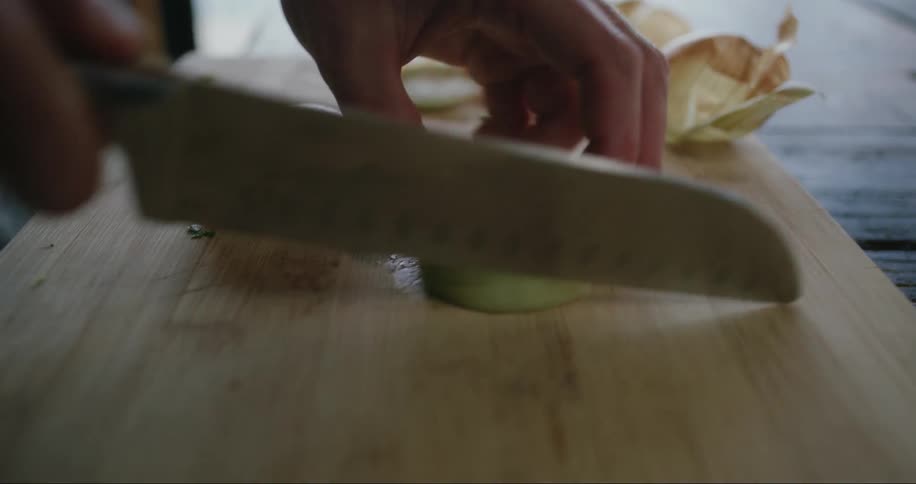Close up of a person using a knife to chop an onion on wooden cutting board as daylight shines through