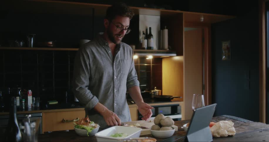 Man cutting and preparing food in his kitchen while talking to someone on the laptop in front of him