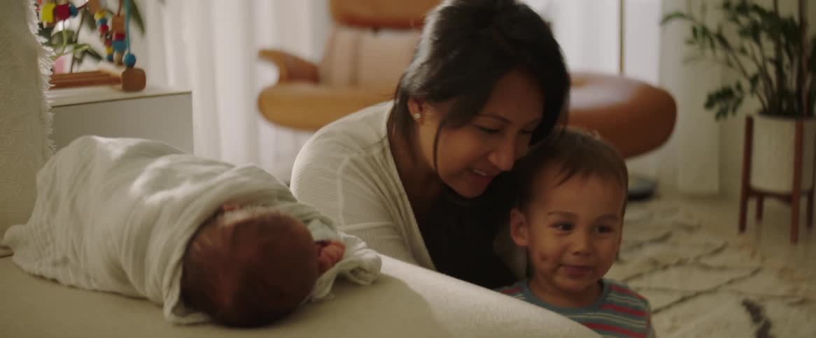 Mother playing with her eldest son on the floor and her baby sleeping on the sofa