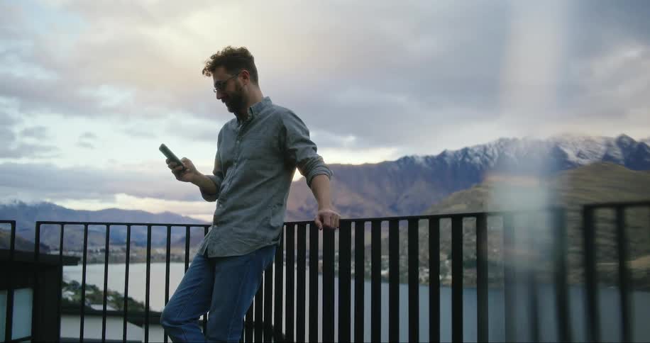 A man with glasses is operating his smartphone while leaning beside a handrail close to a river next to a mountain