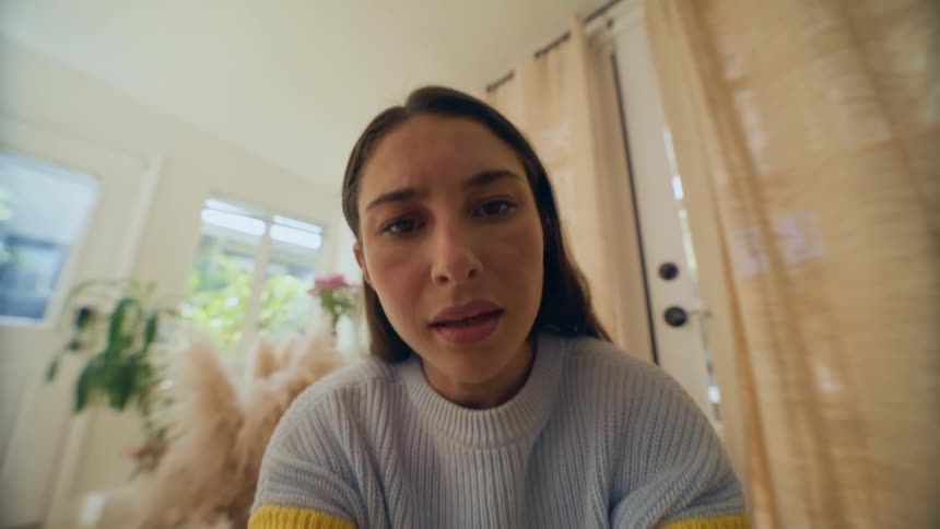 Seated woman picks up the camera that is on the floor brings it to her face says something and puts the camera back on the floor
