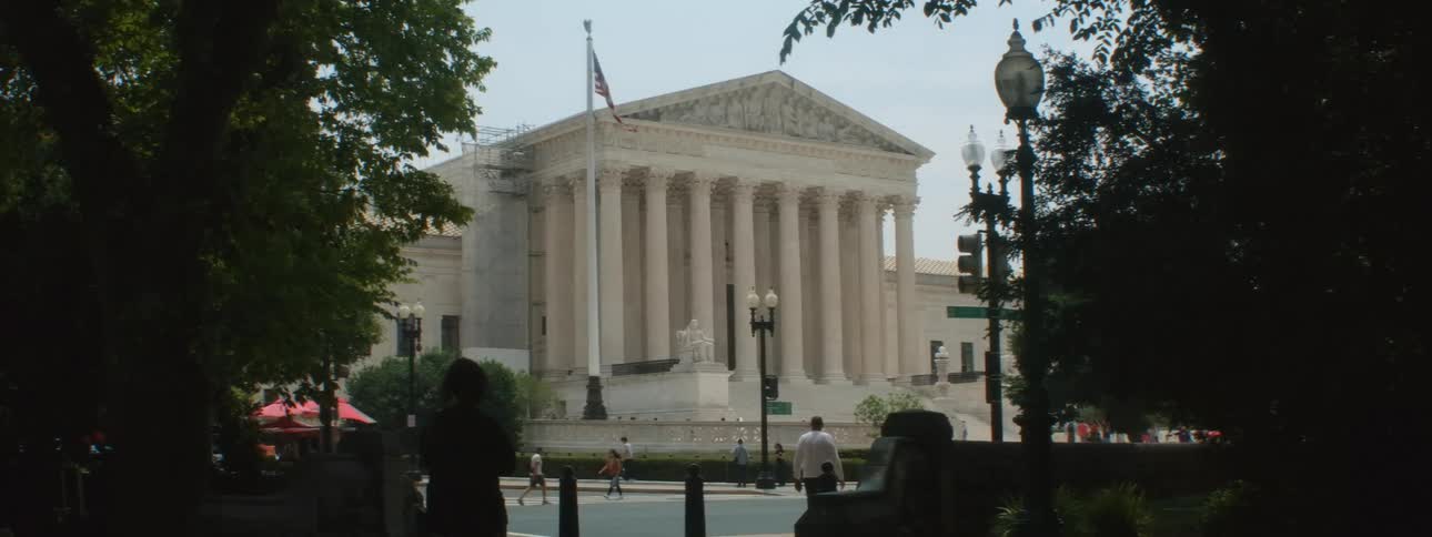 A building of ancient architecture with huge pillars in front of a sidewalk where pedestrians are walking