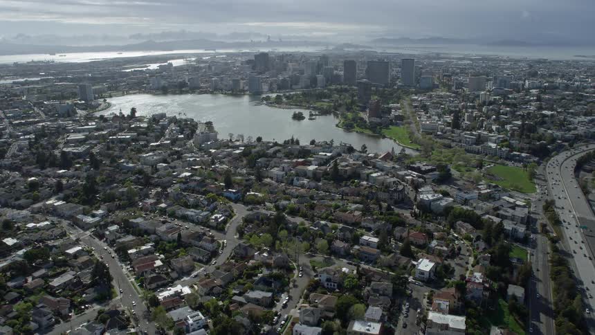 Aerial footage of urban neighborhoods in Oakland California showcasing the approach to Lake Merritt and downtown with partly cloudy skies and wide-angle views from a shotover f1 camera