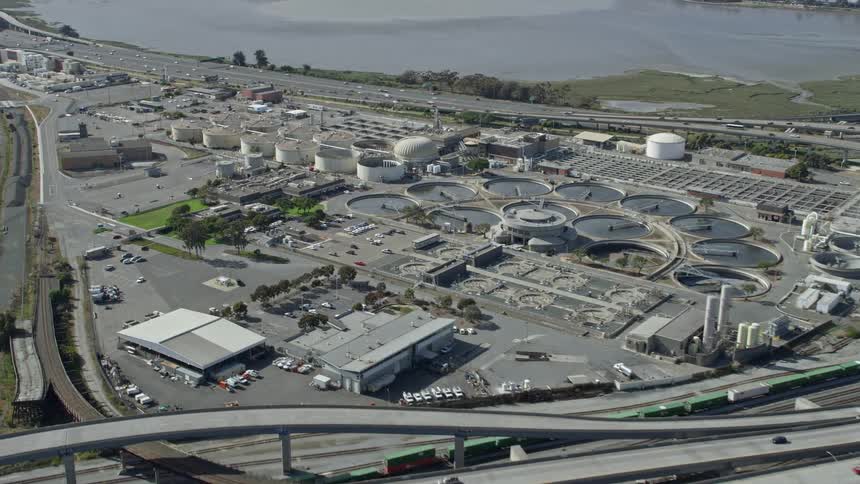  aerial footage of the East Bay Municipal Utility District wastewater treatment plant in Oakland California on a sunny day showcasing the industrial atmosphere and the city landscape surrounding it