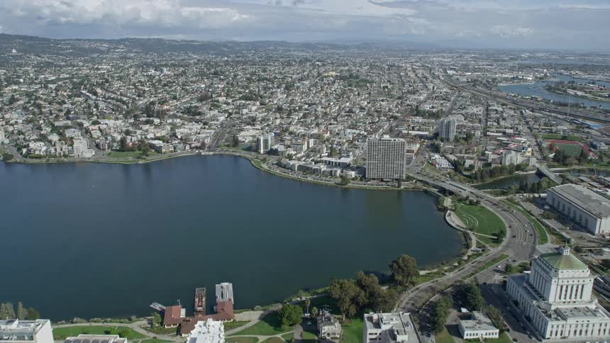  aerial footage capturing an approach to urban neighborhoods in Oakland California flying over Lake Merritt with partly cloudy skies and a wide-angle view of the city's downtown area during the daytime