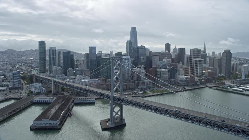 Aerial footage of aclip flying over the Bay Bridge towards the downtown San Francisco skyline with high rises and skyscrapers in view showcasing the cityscape and transportation in Northern California