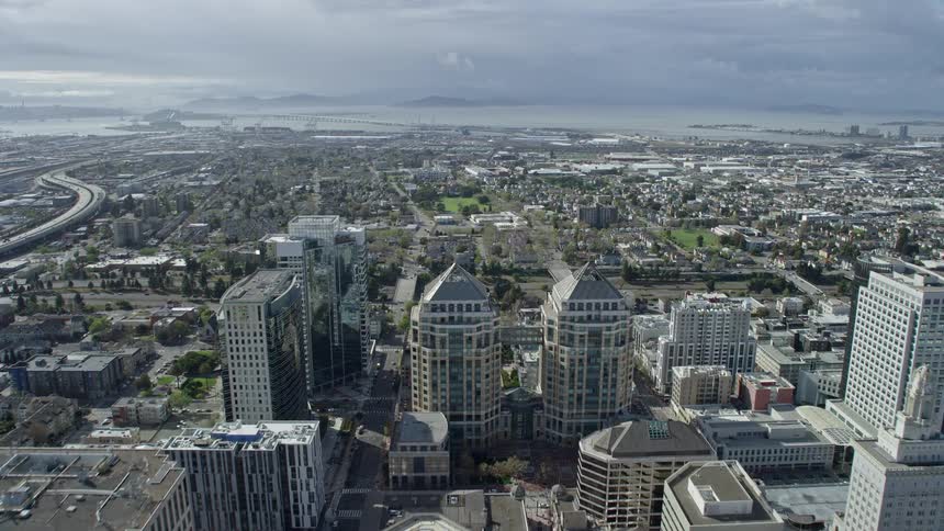 Aerial footage of flying over Tribune Tower in Downtown Oakland towards urban neighborhoods in California during the day showcasing office buildings industry and residential areas with partly cloudy skies