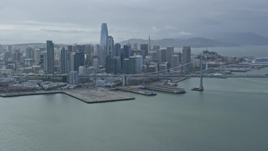  aerial footage showcasing the stunning skyline of Downtown San Francisco California as seen from the bay featuring high-rise buildings the iconic Bay Bridge and cloudy skies during daytime