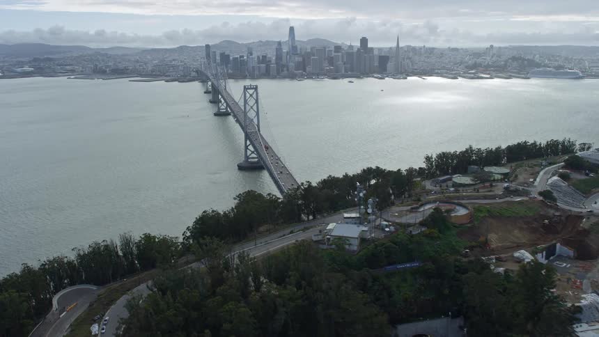  aerial footage of the Bay Bridge tilt up to showcase the stunning downtown San Francisco skyline over the San Francisco Bay capturing the cityscape suspension bridge cloudy skies and light traffic