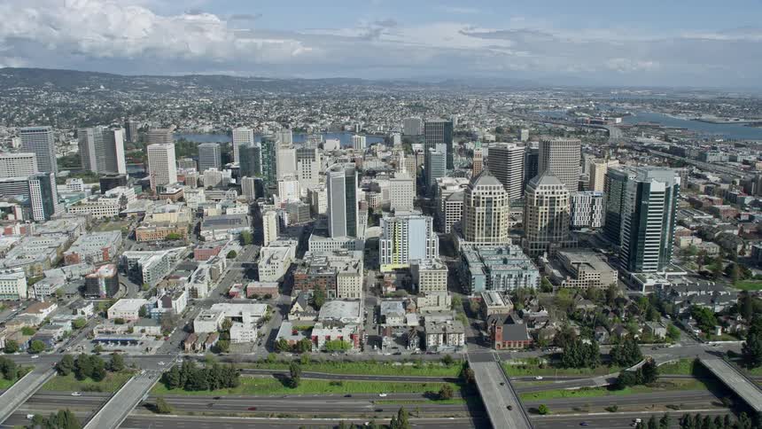  aerial footage showcasing the vibrant cityscape of Downtown Oakland as we soar over office buildings towards the beautiful Lake Merritt on a partly cloudy day in Northern California's Bay Area
