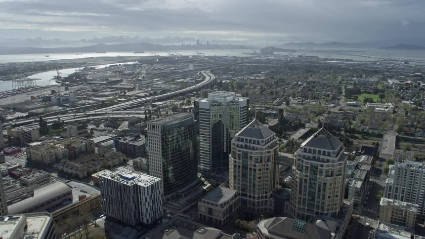 Aerial footage of downtown office buildings and an interstate highway as it approaches the Port of Oakland California with cloudy skies and a view of the river in the background