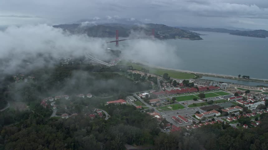  aerial footage of office buildings and studios at The Presidio in San Francisco California with a wide-angle view of the iconic Golden Gate Bridge surrounded by foggy and cloudy skies during the day