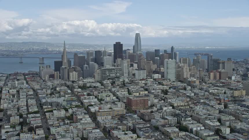  aerial footage showcasing the expansive cityscape and skyline of Downtown San Francisco California with high-rise buildings towers and skyscrapers against a cloudy backdrop capturing a wide-angle side view of the city's superstructures in the spring/summ