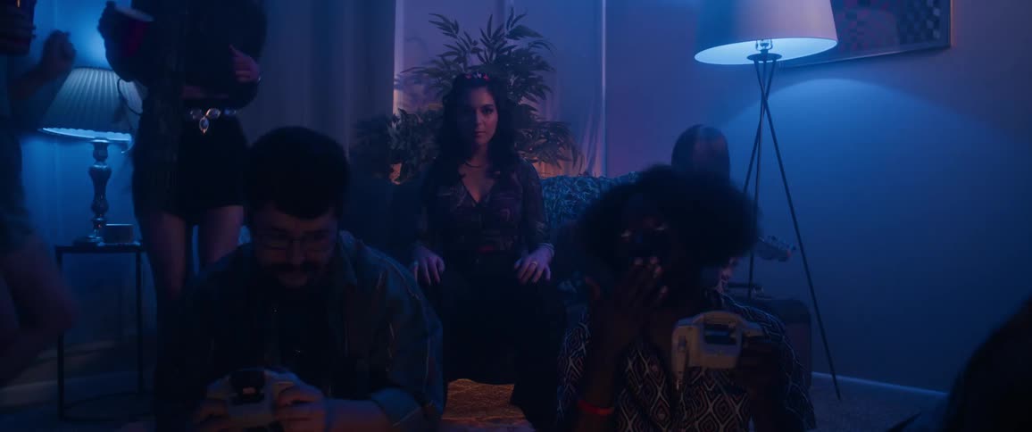 In a dimly lit room a group of friends gathers together two of them sit on a couch one holding a guitar while a standing lamp illuminates the scene an art piece hangs on the wall behind the couch