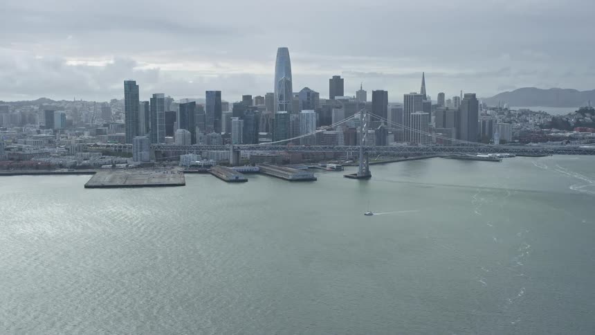 A stunningaerial footage showcasing the city skyline of Downtown San Francisco California with the iconic Bay Bridge and boats passing underneath on a cloudy day