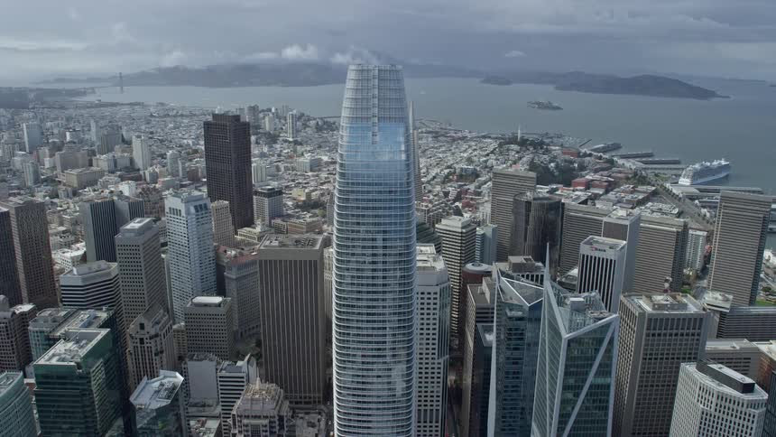 Aerial footage flies over Salesforce Tower towards Transamerica Pyramid in downtown San Francisco showcasing the iconic structures and city skyline on a cloudy day