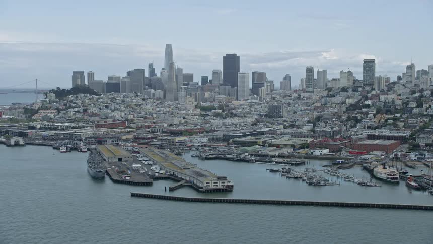  aerial footage of the iconic San Francisco skyline viewed from Fisherman's Wharf showcasing the city's high-rise buildings waterfront boat docks and partly cloudy skies during a sunny day