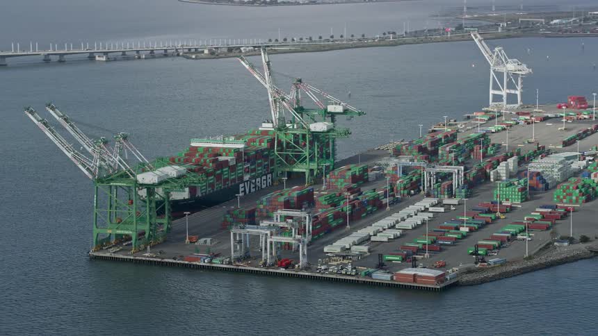  aerial footage of cargo cranes and a ship passing by at the Port of Oakland in California during the daytime showcasing the industrial and maritime atmosphere of the area