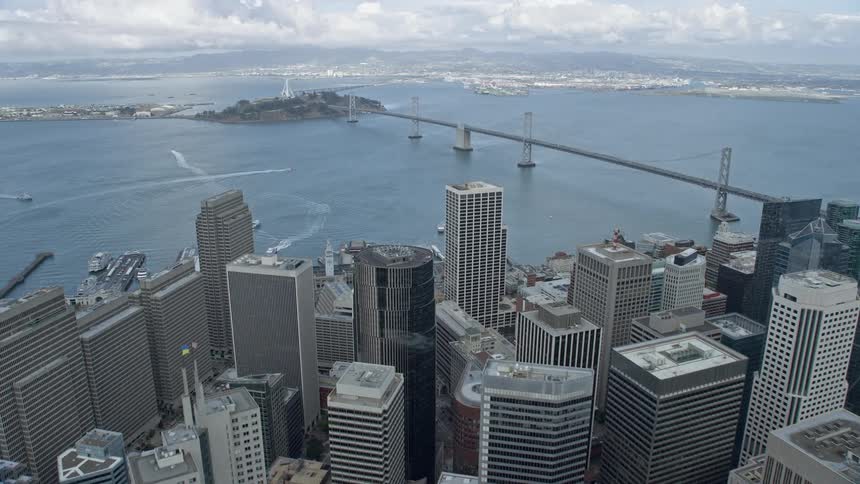  aerial footage captures an flyover of downtown San Francisco showcasing the iconic Bay Bridge and Yerba Buena Island amidst the city's towering skyscrapers and cloudy skies