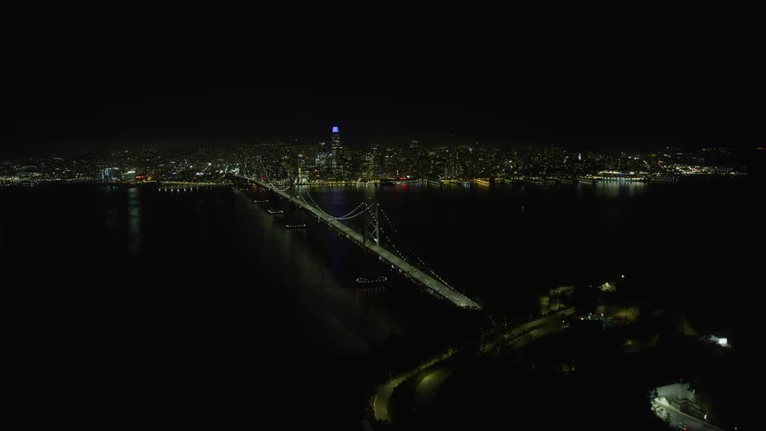 aerial footage ascending over Yerba Buena Island revealing the illuminated Bay Bridge and Downtown San Francisco at night in California with a clear night sky