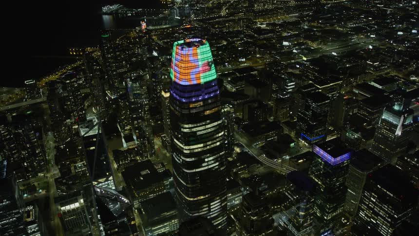  aerial flyby of Salesforce Tower at night in Downtown San Francisco California showcasing the city skyline's colorful lights and clear skies