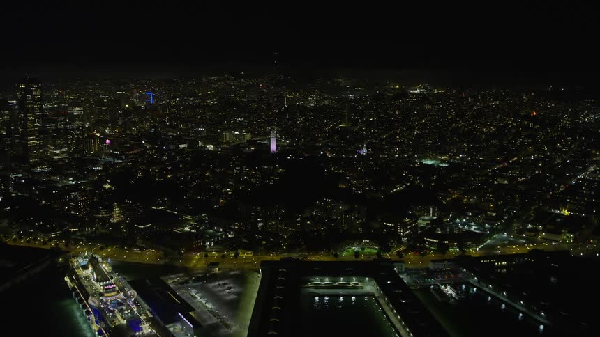 aerial footage of Coit Tower at night in San Francisco California showcasing the city's majestic skyline illuminated by sparkling lights and a docked ship emanating a radiant glow