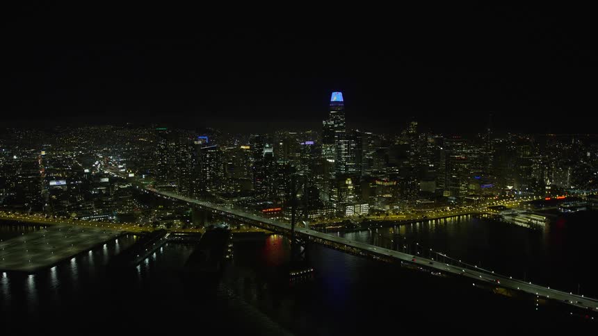  aerial footage of a clear night flyover of Bay Bridge approaching Downtown San Francisco skyline with brightly lit high-rise buildings and a reflection of the bridge on the water surface