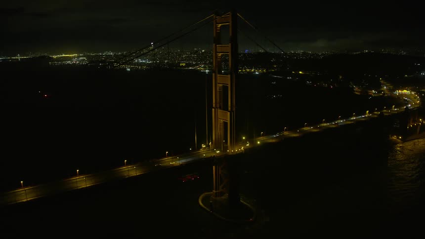  aerial footage of the iconic Golden Gate Bridge at night with San Francisco in the background showcasing the historic landmark and light traffic in clear skies shot from a side view perspective above Marin County California