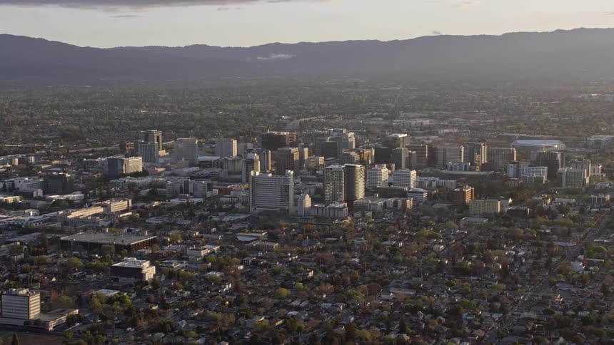 Breathtakingaerial footage showcasing the cityscape of Downtown San Jose at dusk with a hazy sunset backdrop highlighting the vibrant downtown area and its surrounding suburban neighborhoods set against the majestic mountains in the horizon