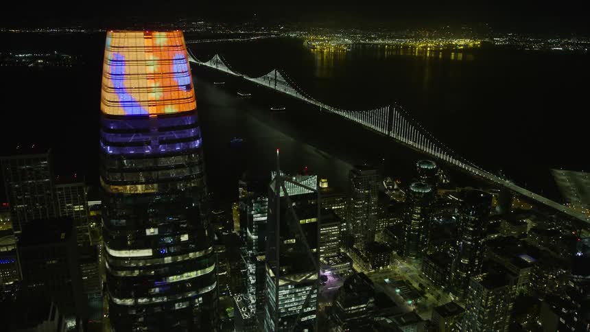  aerial footage showcasing an nighttime view of San Francisco's downtown flying towards the illuminated Bay Bridge surrounded by clear skies and towering skyscrapers in the vibrant city of northern California