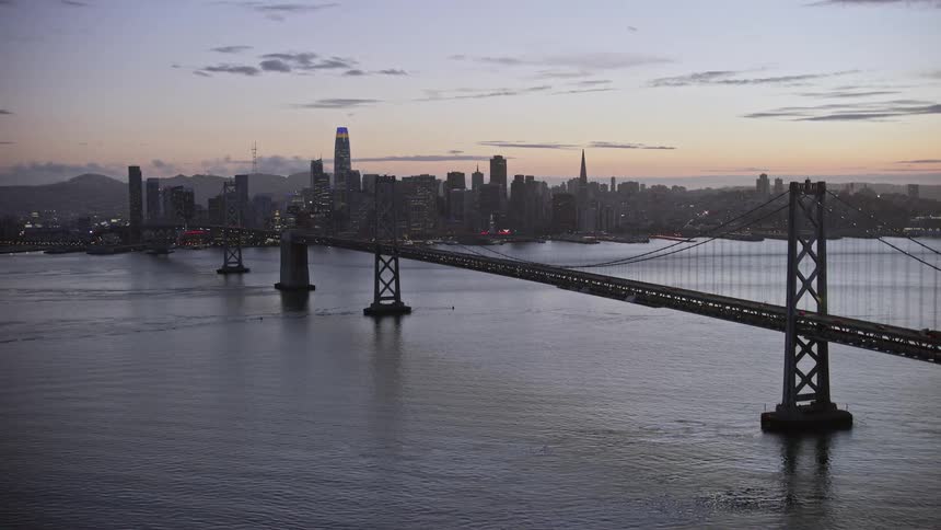  aerial footage captures the enchanting twilight scene of Downtown San Francisco skyline and Bay Bridge connecting the metropolitan city over water during a partly cloudy summer showcasing the high rises skyscrapers and superstructures of this iconic Cali
