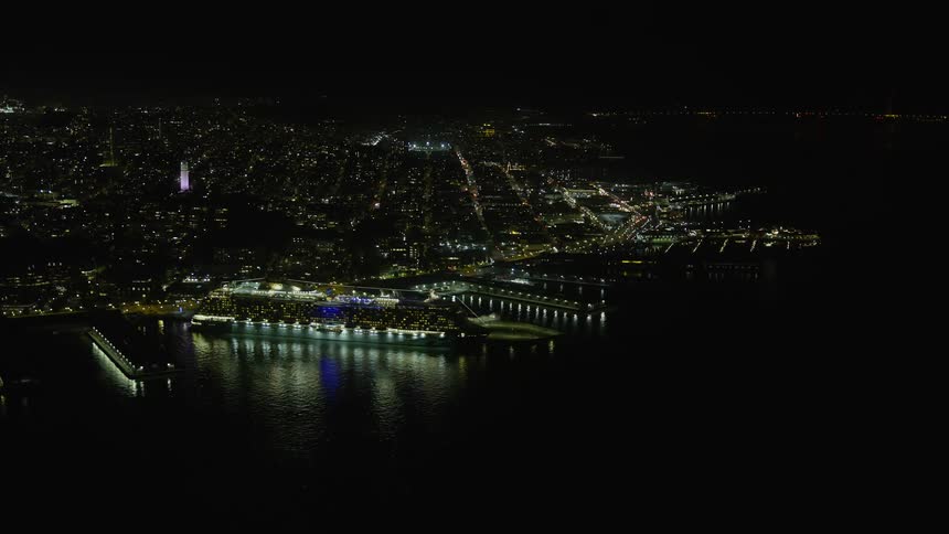 an aerial view of a large cruise ship illuminated at night docked near Coit Tower in San Francisco California