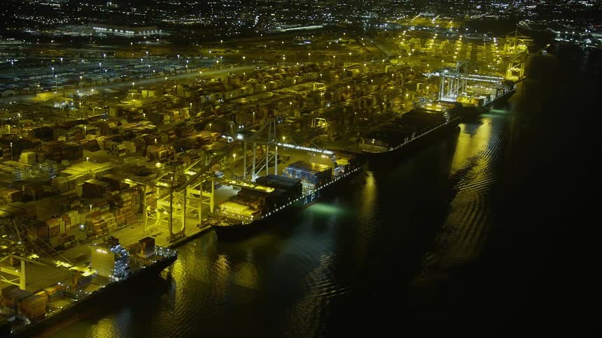 Aerial footage at night of cargo ships docked at the Port of Oakland California with bright lights illuminating the container yard and a line of cranes along the bay