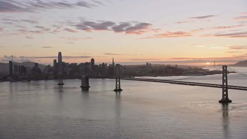  aerial footage of the Bay Bridge and the Downtown San Francisco skyline at sunset showcasing the high rises cityscape and the partly cloudy skies over the magnificent suspension bridge and towers capturing the essence of the city's evening beauty in Nort