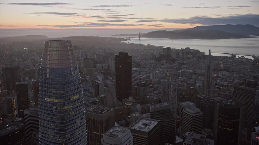 aerial footage capturing the mesmerizing cityscape of Downtown San Francisco at twilight as the camera flies over the bustling downtown area towards the iconic Golden Gate Bridge in the distance against a backdrop of partly cloudy skies and the beautiful