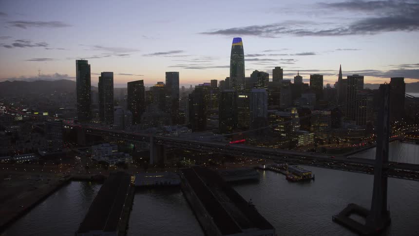 Aaerial flyby of the Bay Bridge at twilight showcasing the downtown San Francisco skyline with high-rise buildings and skyscrapers overlooking the cityscape of Northern California as seen from the scenic Bay Area on the west coast