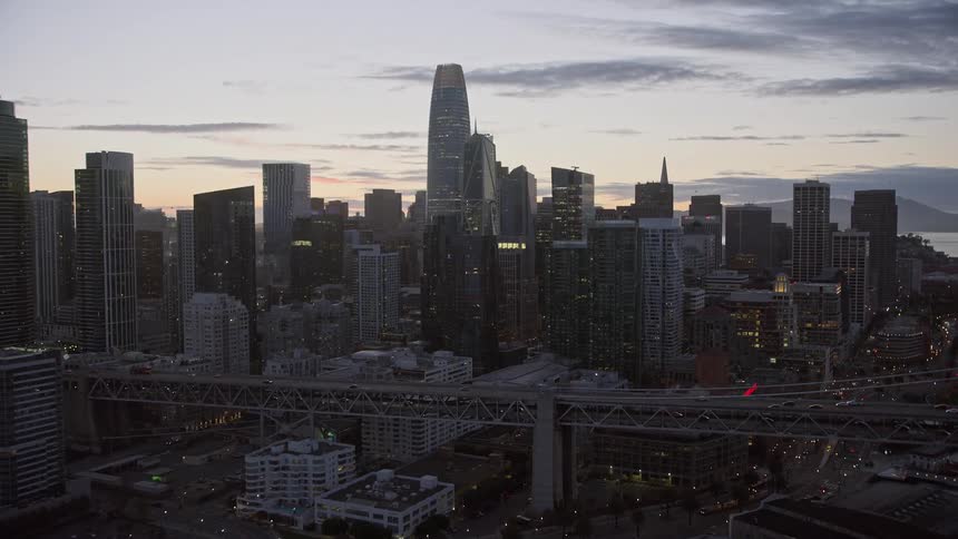  aerial footage showcasing the stunning cityscape of downtown San Francisco at twilight as the camera ascends towards the high-rise buildings and towering skyscrapers approaching from the bay area with the Bay Bridge in view capturing the essence of the b