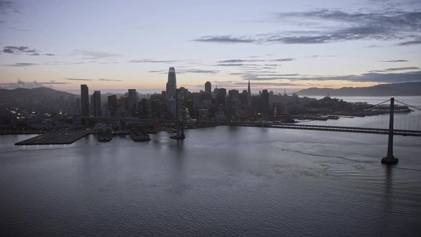 A stunningaerial view of the Downtown San Francisco skyline at twilight showcasing the high rises skyscrapers and the iconic Bay Bridge as the aerial footage flies away from the city capturing the beauty of Northern California's west coast