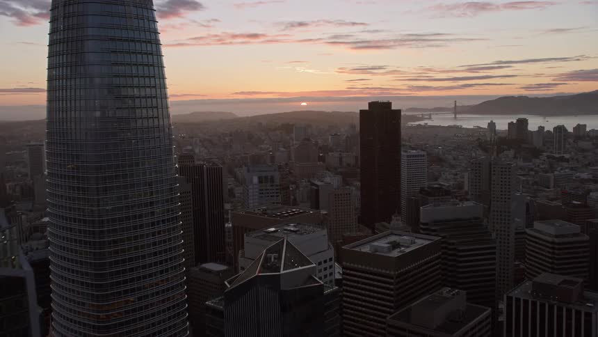 Drone flies over Downtown San Francisco at sunset capturing the city skyline Bay Bridge and partly cloudy skies