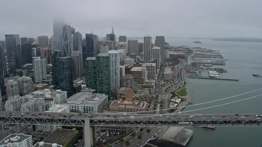  aerial footage of a foggy day in San Francisco flying over the Bay Bridge and past Downtown with high rise buildings and skyscrapers showcasing the city's downtown area and the large cable-stayed bridge over the San Francisco Bay