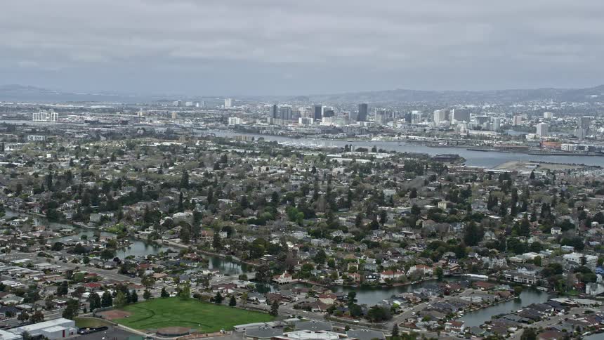  aerial footage captures the suburban landscape of Alameda California with a cloudy sky showcasing the residential neighborhoods and downtown areas of Oakland in the backdrop