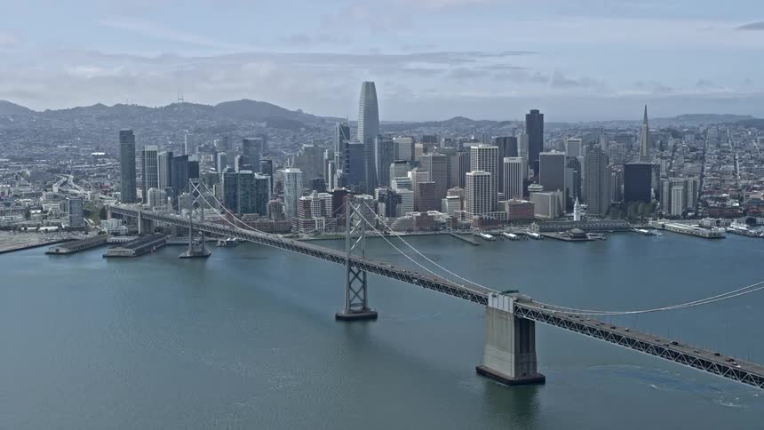 Aerial footage of Downtown San Francisco skyline seen from near the Bay Bridge with traffic crossing the suspension bridge towards the city on a partly cloudy day in California during summer