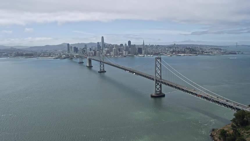  aerial footage showcasing the ascend over Yerba Buena Island revealing the Bay Bridge and Downtown San Francisco on a partly cloudy day in Northern California