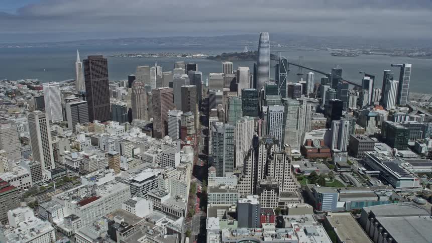 Aerial footage of a summer day in downtown San Francisco California showing high rise buildings and city streets as it follows Market Street towards downtown with a wide-angle view of the city and partly cloudy skies