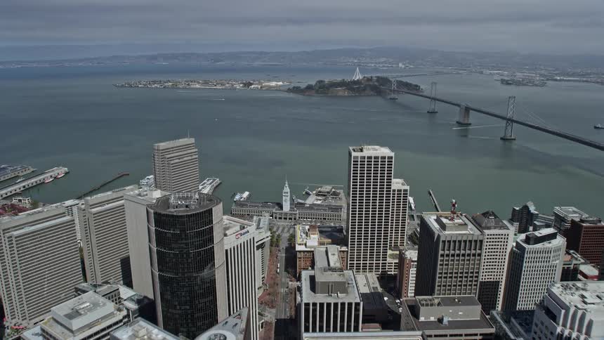 Aerial footage of Downtown San Francisco during the day flying over Market Street and approaching the bay with partly cloudy skies showcasing high-rise buildings and the iconic Bay Bridge connecting the city to another urban area